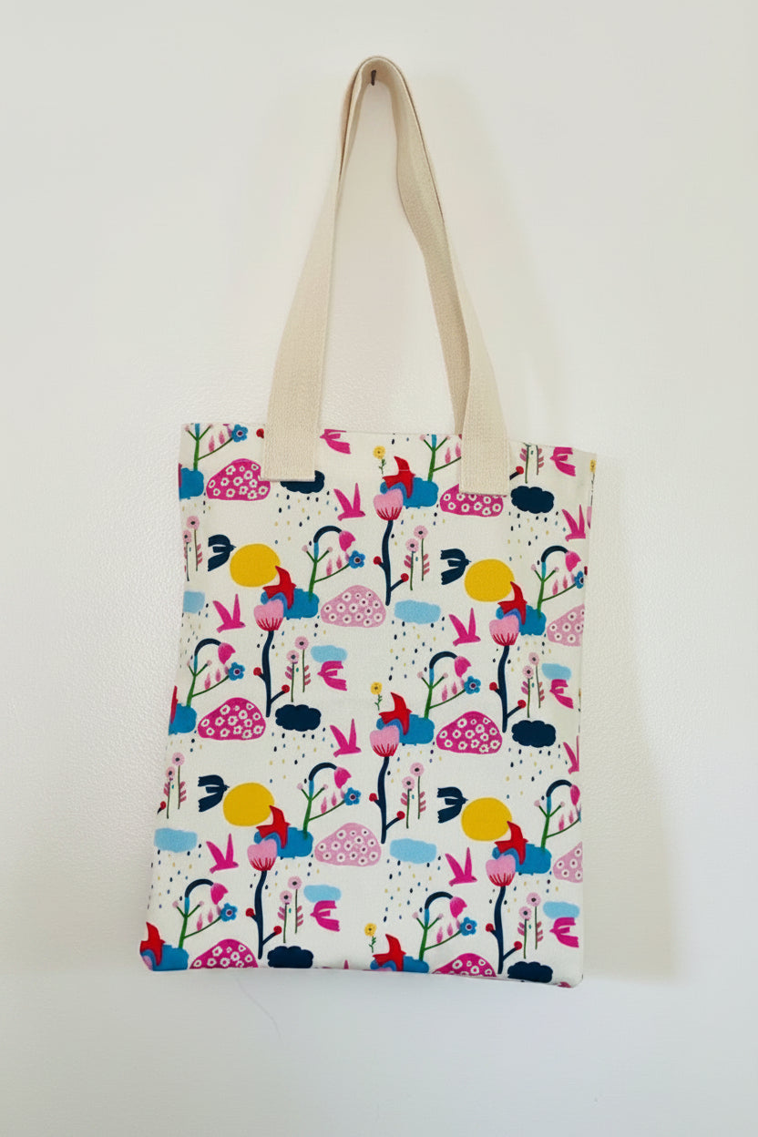 A canvas tote bag with a colorful, whimsical folk-art style pattern of birds, flowers, and mushrooms on a cream background, featuring natural beige shoulder straps.